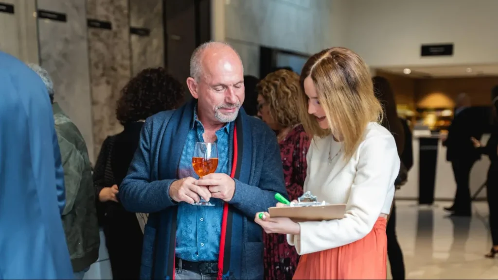 A man and woman networking at a corporate event, holding a clipboard and a glass of wine.