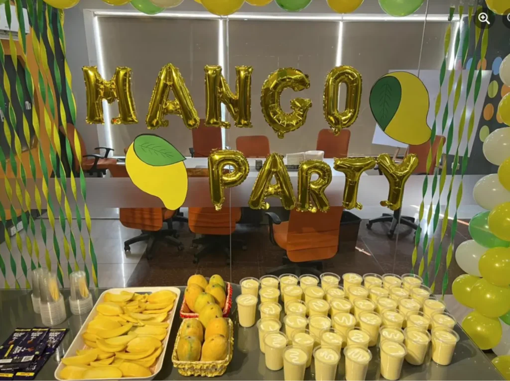 Decorated table with mangoes, mango shakes, and golden 'Mango Party' balloons at a corporate event.