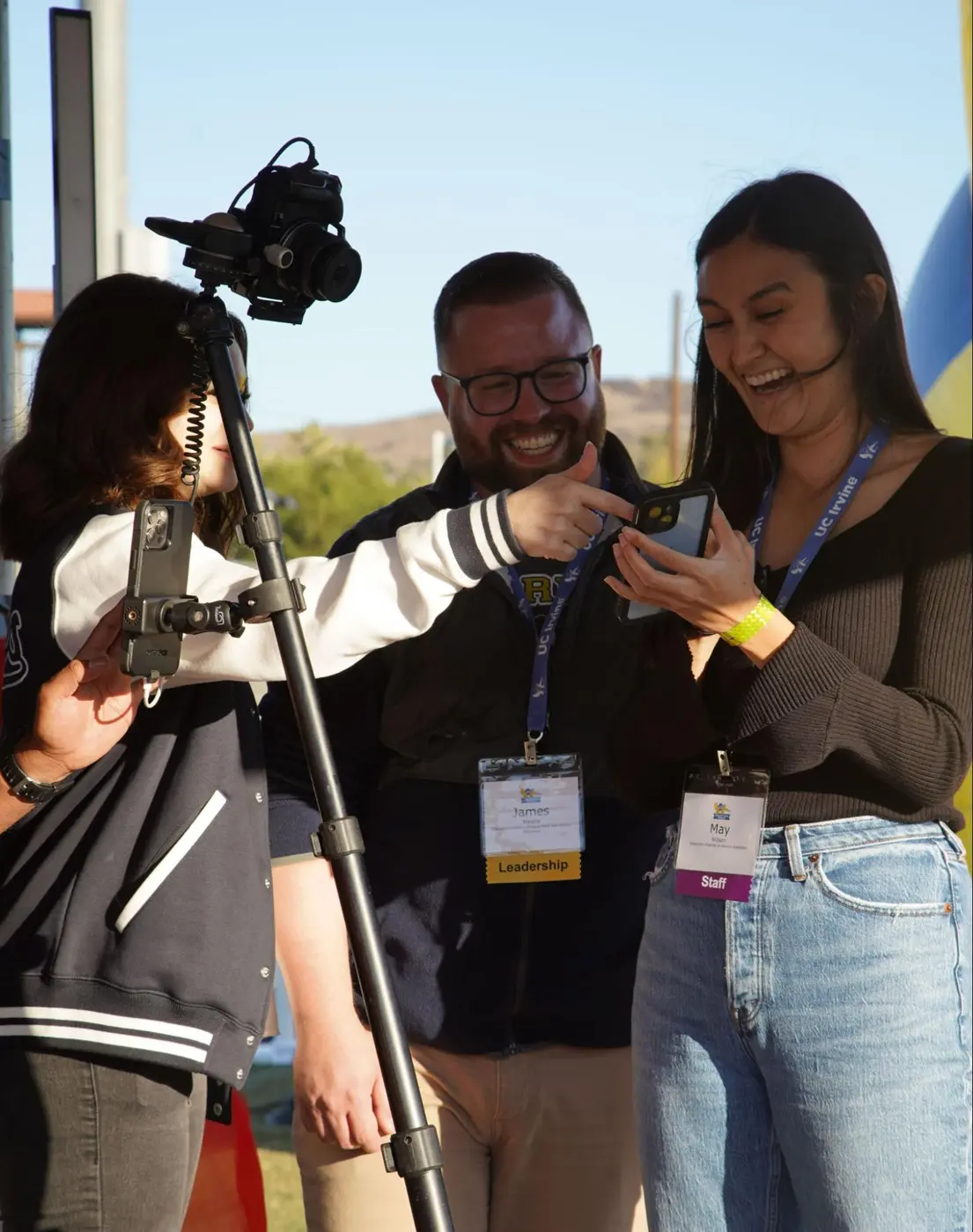 Guests laughing while reviewing a video confessional recording on a phone at an outdoor event setup.