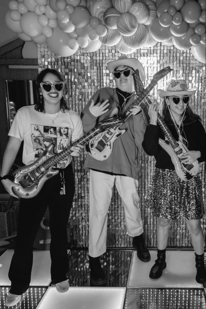 Three people in heart-shaped sunglasses posing with inflatable guitars at a fun corporate party.
