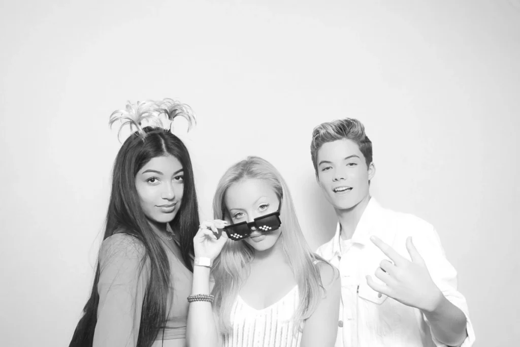 Black and white photo booth portrait of three young friends posing playfully with pixel sunglasses and headpiece