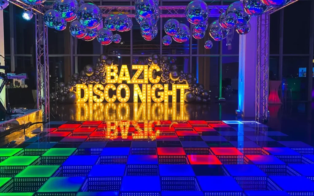 LED dance floor with multicolored panels beneath 'BAZIC DISCO NIGHT' marquee sign and mirror balls, showcasing event theming