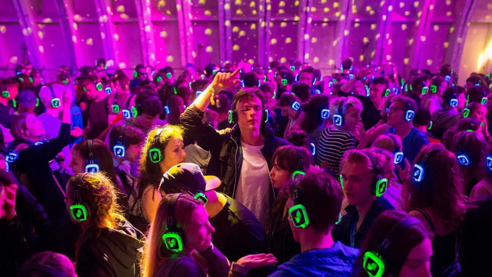 Packed silent disco dance floor with crowd wearing synchronized green glowing headphones under purple lighting