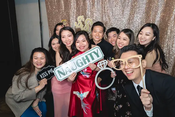 Wedding photo booth group shot with props including 'Naughty' sign and oversized ring frame against sequin backdrop