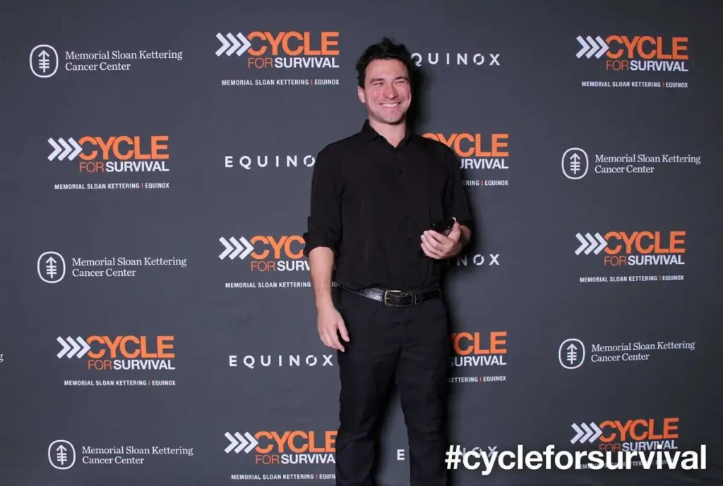 Participant posing at Cycle for Survival charity event with branded step-and-repeat backdrop and event hashtag