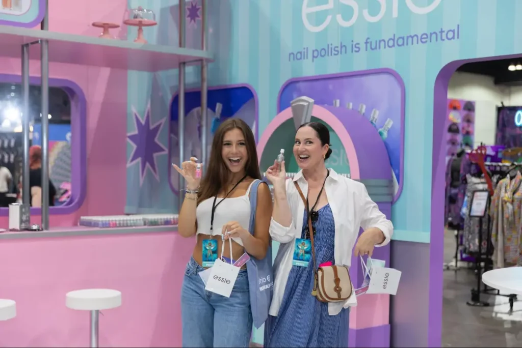 Two women at an Essie nail polish promotional event, holding sample products and shopping bags in a colorful branded booth