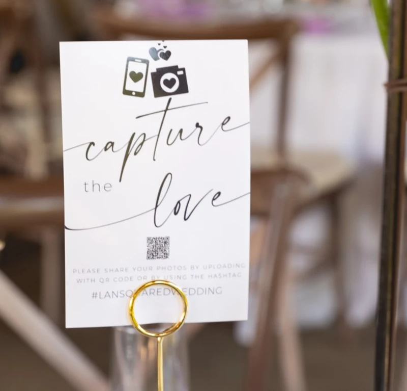 Wedding table card with 'capture the love' text, smartphone and camera icons, QR code, and hashtag for photo sharing