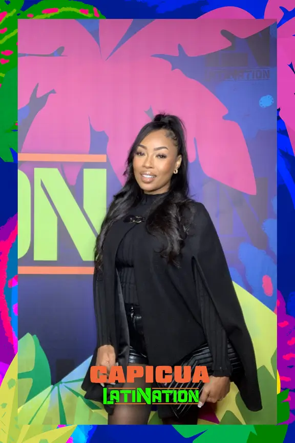 Woman in black outfit posing in front of vibrant pink digital backdrop with CAPICUA LATINATION branding, using AI photo frame