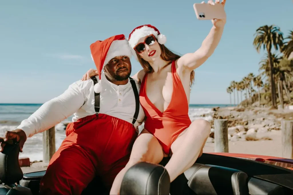 Couple in festive Santa hats and red outfits taking selfie at beach, ideal for tropical holiday-themed parties