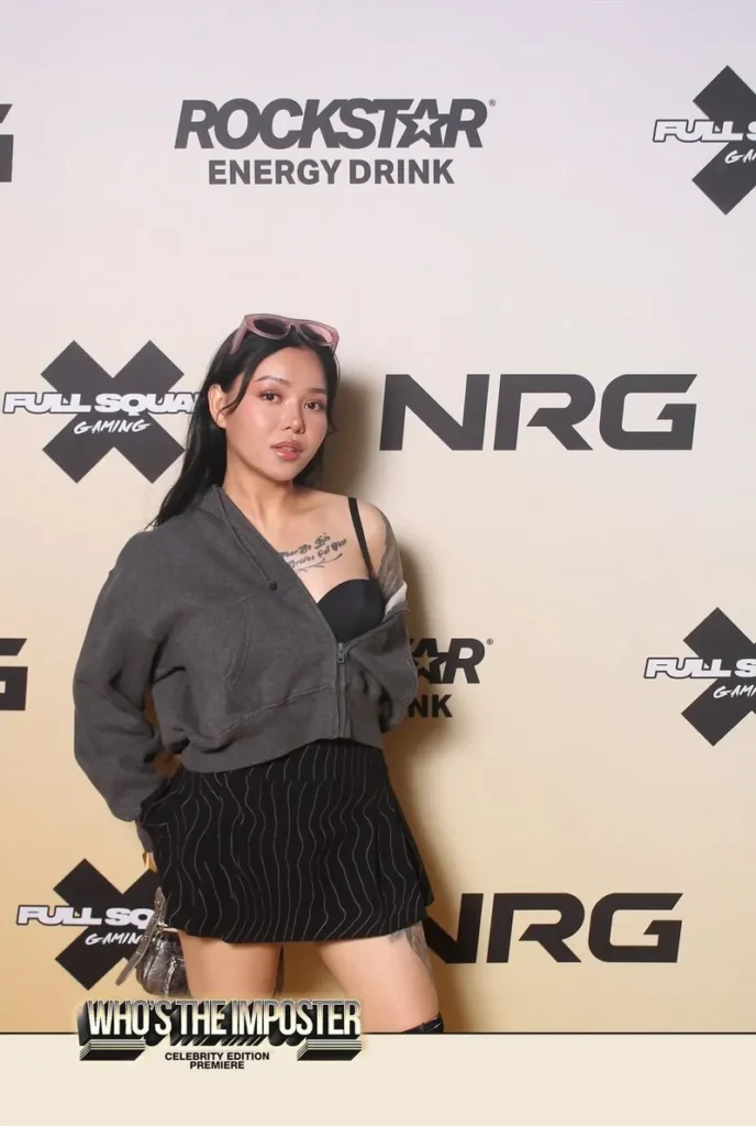 Person posing at gaming event with Rockstar Energy and NRG sponsor backdrop for 'Who's The Imposter' celebrity edition