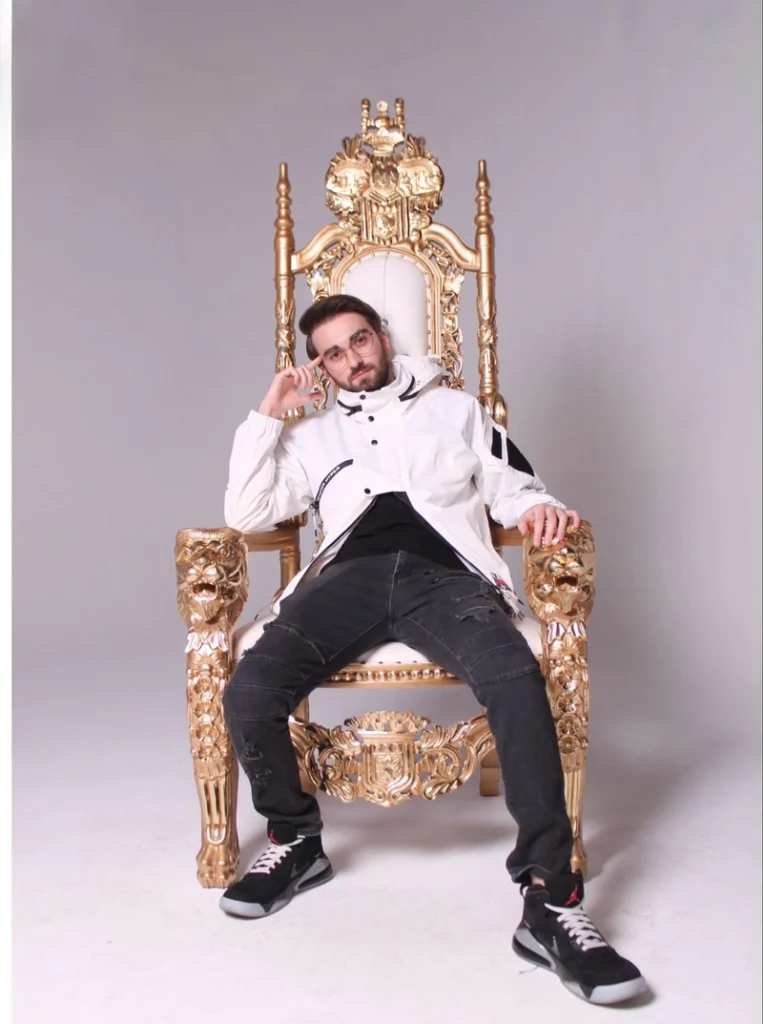 Person seated on ornate gold throne wearing white jacket and dark jeans in professional photo studio setting