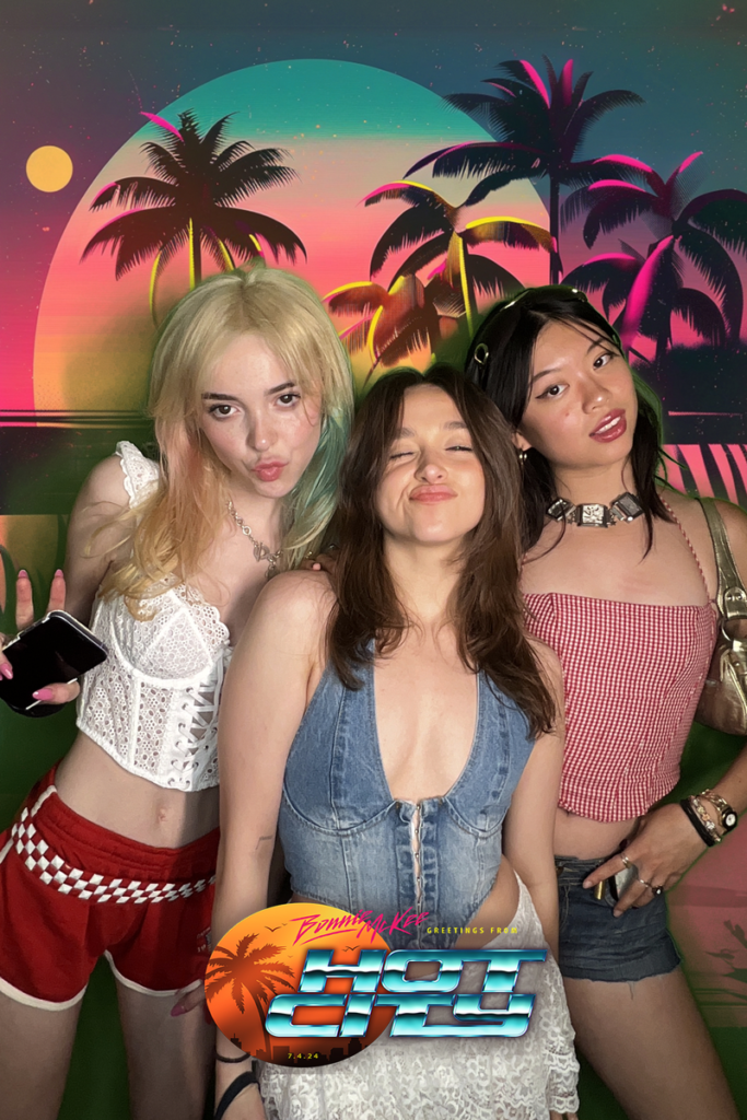 Three women posing in front of a tropical backdrop with neon palm trees and 'HOT CITY' text - Miami vice themed party