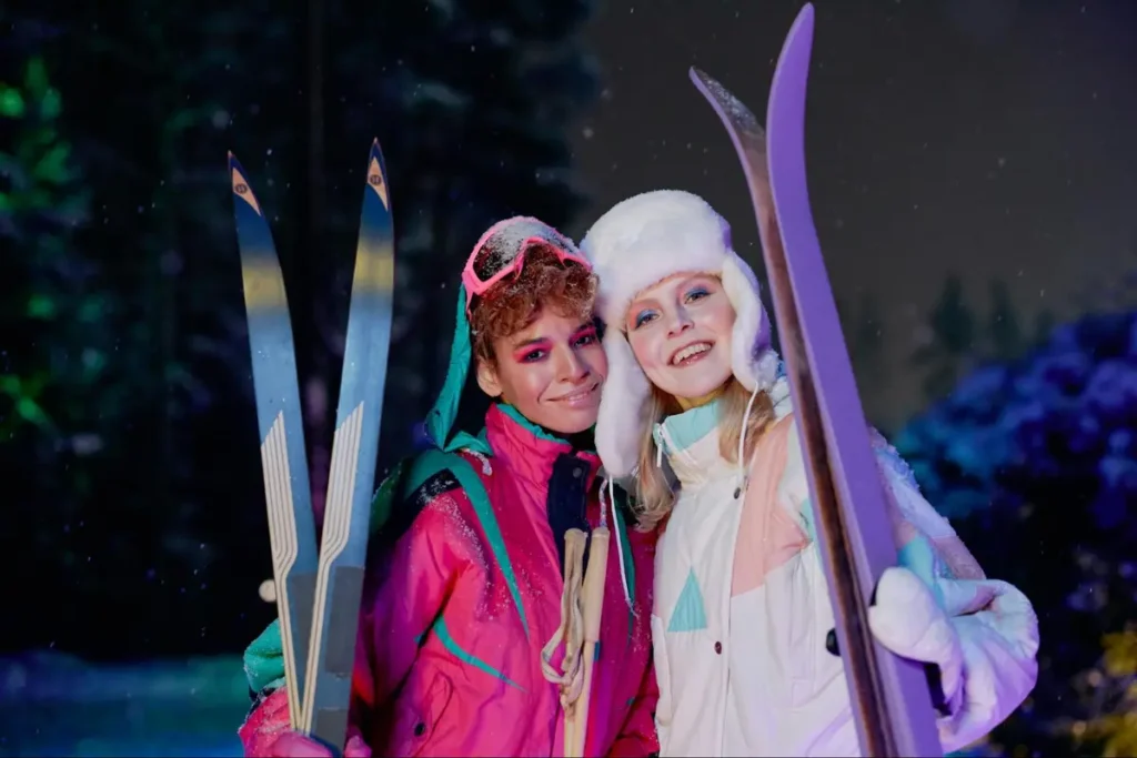 Two friends in colorful 80s-style ski outfits posing with skis against a snowy nighttime backdrop