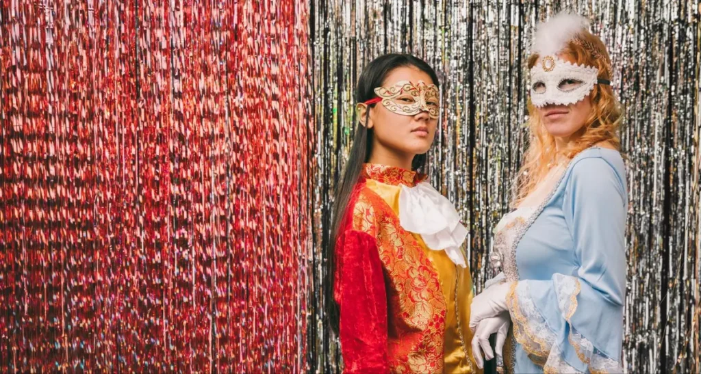 Two women in masquerade attire with ornate masks against red and silver backdrop - elegant masquerade ball theme idea